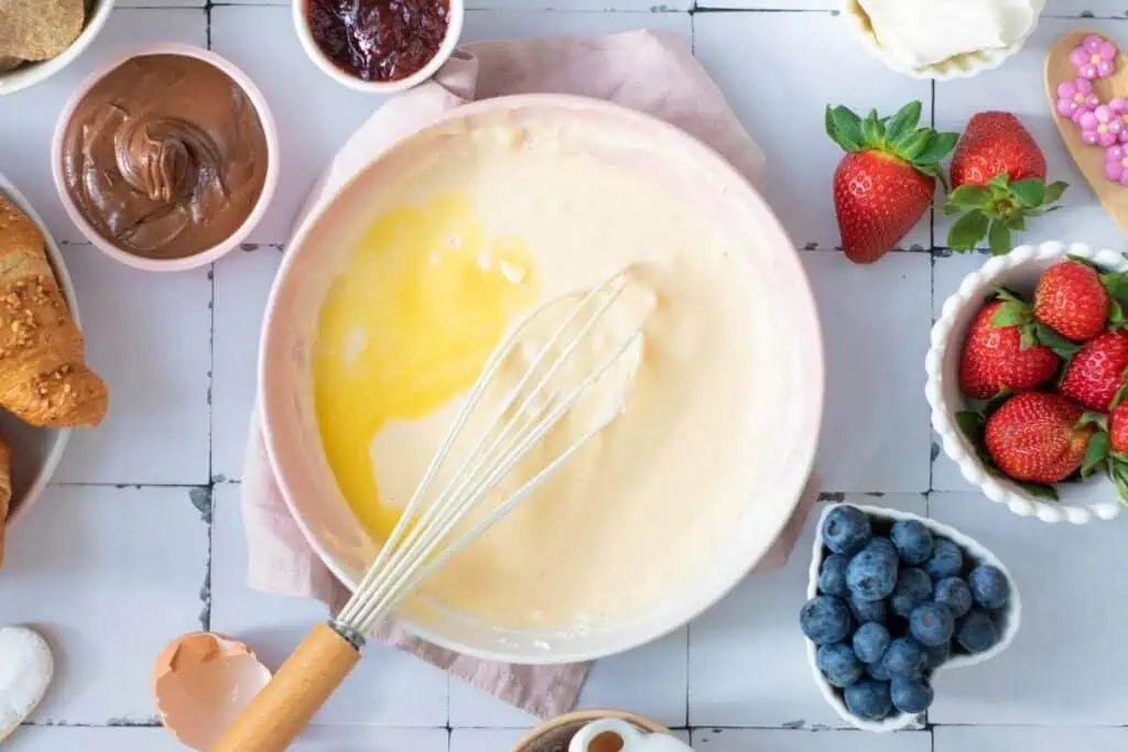 A mixing bowl with pancake batter and a whisk, surrounded by bowls of strawberries, blueberries, chocolate spread, jam, and other breakfast ingredients on a white tiled surface.