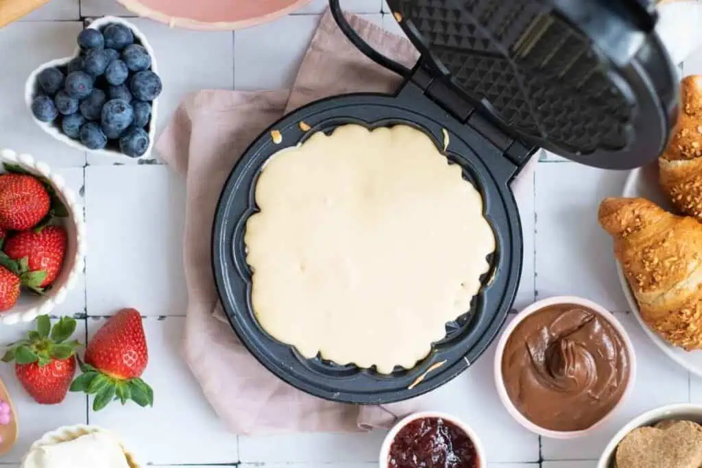 Waffle maker filled with batter surrounded by bowls of blueberries, strawberries, chocolate spread, jam, and croissants on a white surface.