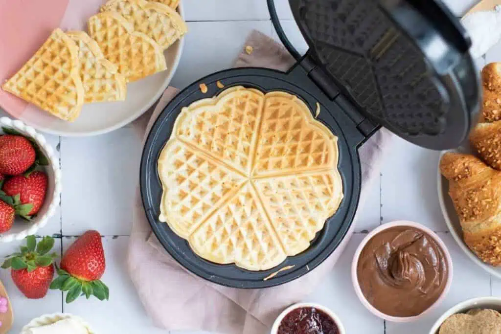Heart-shaped waffles cooking in a waffle maker, surrounded by fresh strawberries, croissants, and bowls of chocolate spread and jam on a white table.