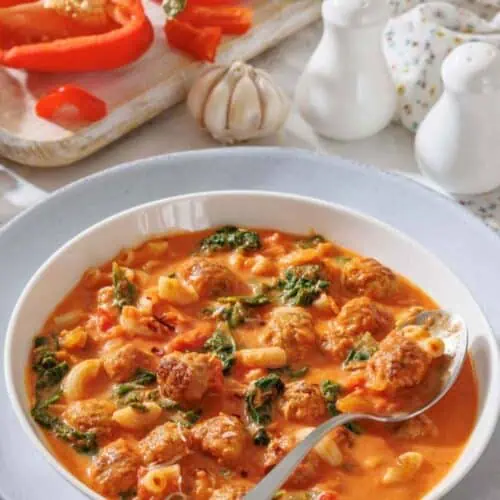 A bowl of soup with meatballs, pasta, spinach, and tomato broth, with a spoon resting inside. In the background are sliced red bell peppers, garlic, and fresh herbs on a cutting board.