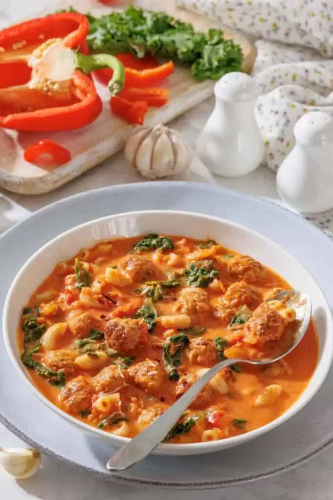 A bowl of soup with meatballs, pasta, spinach, and tomato broth, with a spoon resting inside. In the background are sliced red bell peppers, garlic, and fresh herbs on a cutting board.