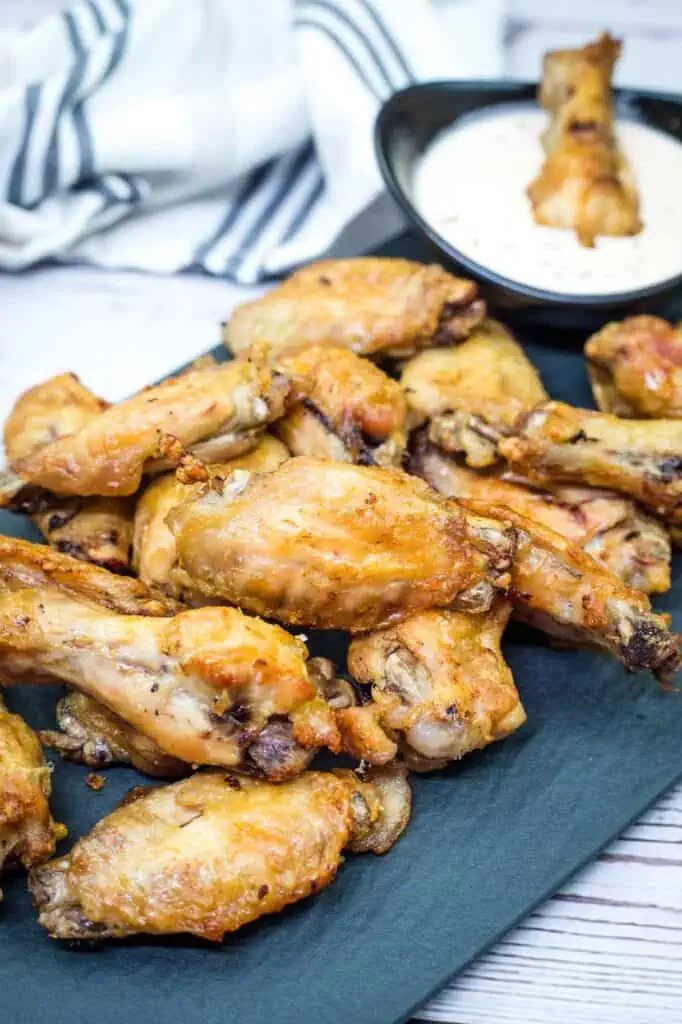 A pile of cooked chicken wings on a dark serving tray with a small bowl of dipping sauce in the background.