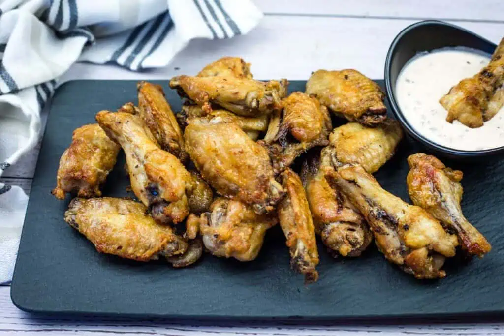 A black serving tray with a pile of cooked chicken wings next to a bowl of creamy dipping sauce, with a striped cloth in the background.