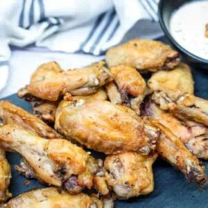 A pile of air fried chicken wings is arranged on a dark serving tray, with a bowl of creamy dipping sauce in the background.