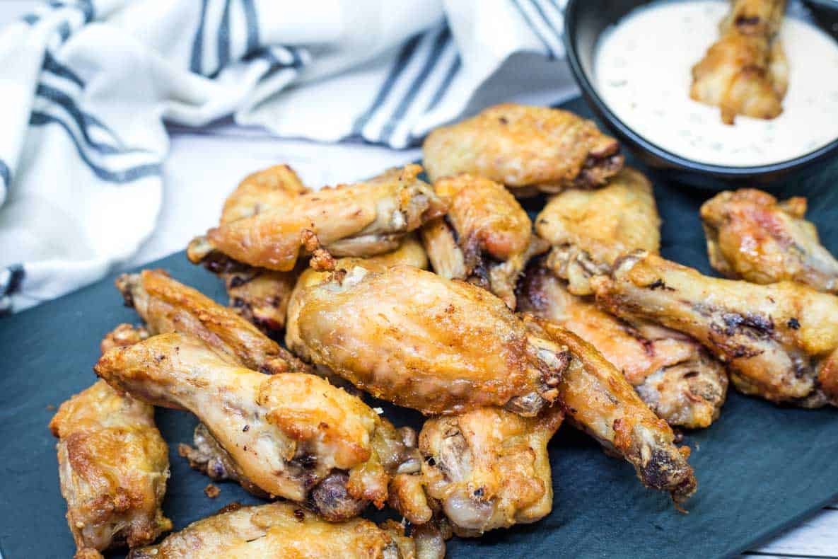 A pile of air fried chicken wings is arranged on a dark serving tray, with a bowl of creamy dipping sauce in the background.