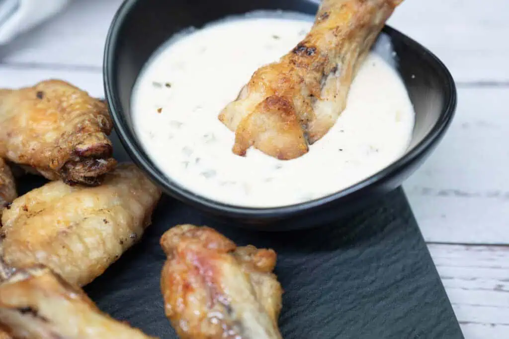 A chicken wing is being dipped into a bowl of creamy white sauce, with more wings placed nearby on a slate surface.