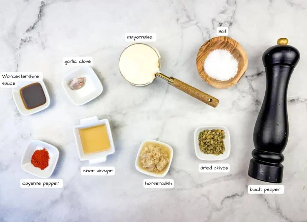 Ingredients for a recipe arranged on a marble surface, including mayonnaise, salt, black pepper, dried chives, horseradish, cider vinegar, cayenne pepper, Worcestershire sauce, and a garlic clove.