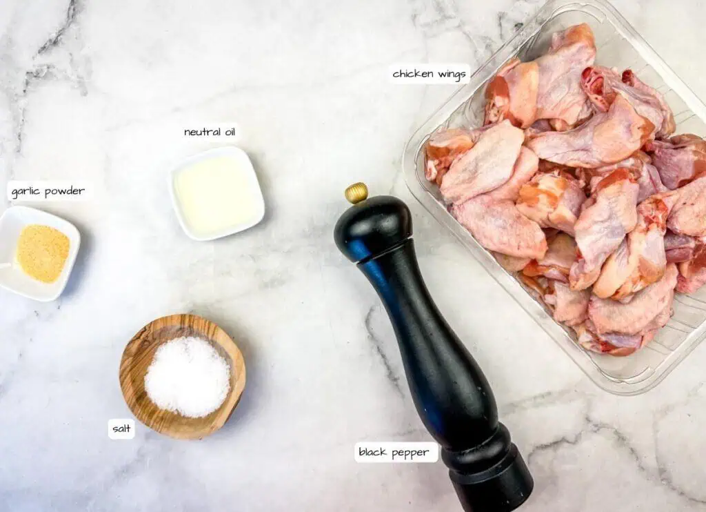 Chicken wings in a plastic container with small bowls of garlic powder, salt, neutral oil, and a black pepper grinder on a marble surface.