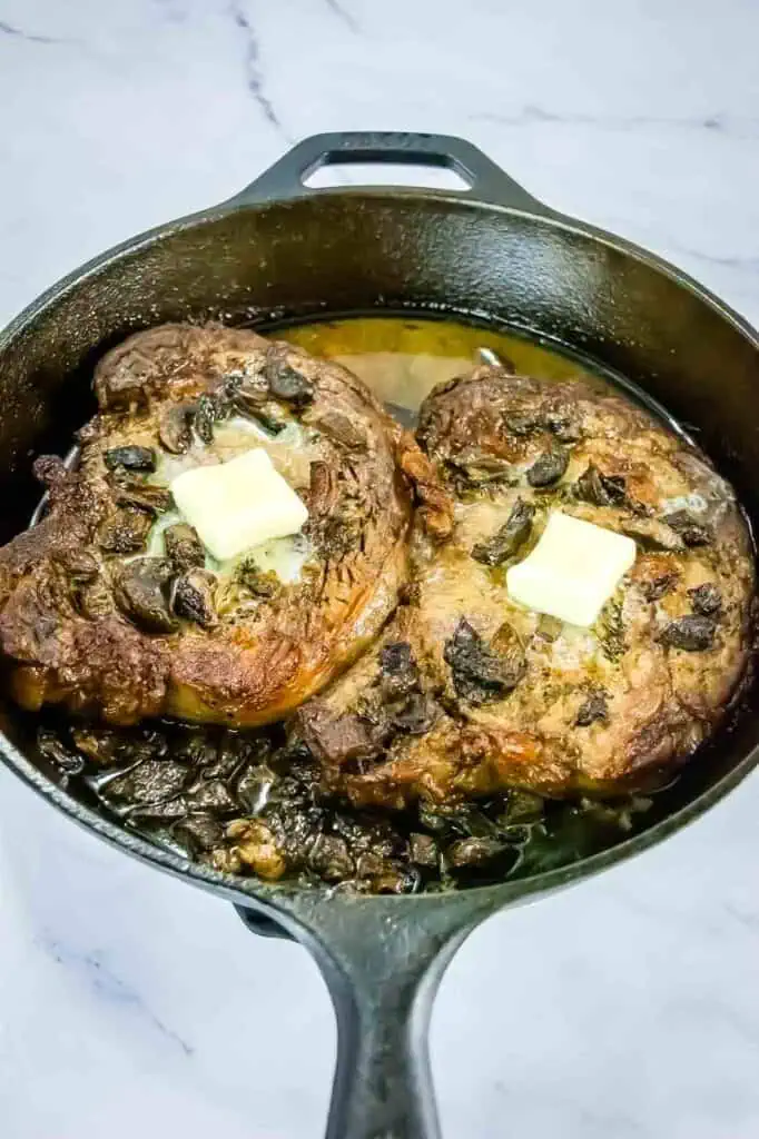 Two cooked steaks with mushrooms and pats of butter on top in a black cast iron skillet on a light-colored surface.