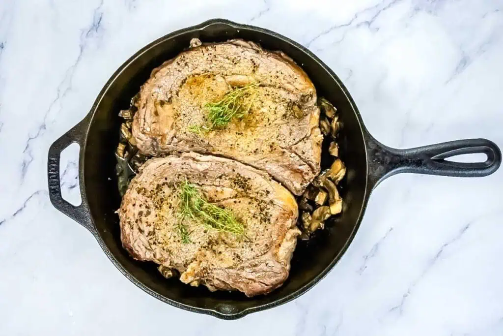 Two cooked ribeye steaks topped with herbs in a cast iron skillet, resting on a bed of chopped mushrooms on a white marble surface.