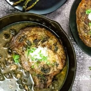 Cast iron skillet with a cooked steak topped with herbs and a dollop of cream, surrounded by a mushroom sauce, with a side plate of steak and asparagus.