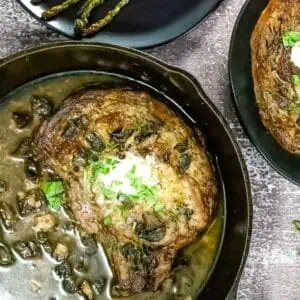 Cast iron skillet with a cooked steak topped with herbs and a dollop of cream, surrounded by a mushroom sauce, with a side plate of steak and asparagus.