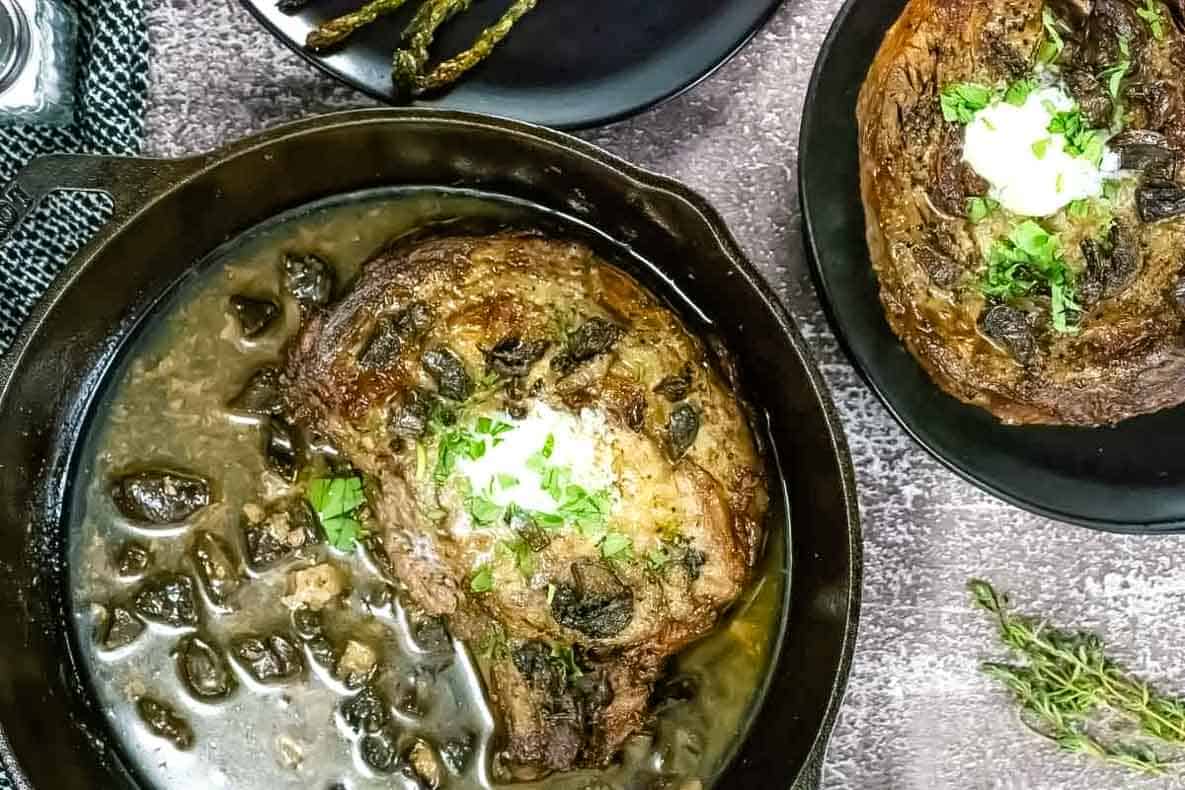 Cast iron skillet with a cooked steak topped with herbs and a dollop of cream, surrounded by a mushroom sauce, with a side plate of steak and asparagus.