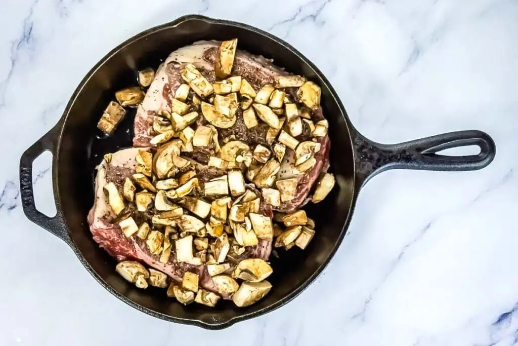 A cast iron skillet containing raw steak pieces topped with chopped mushrooms, placed on a white marble surface.