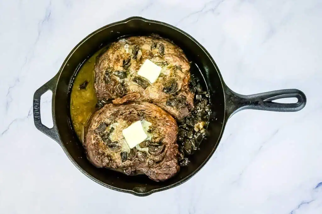Two cooked steaks topped with pats of butter sit in a cast iron skillet with juices, on a white marble surface.