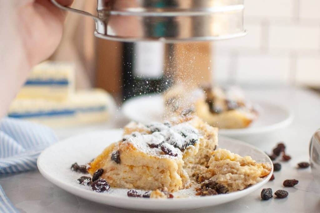 A dusting of powdered sugar is being sprinkled over a slice of bread pudding with raisins on a white plate.