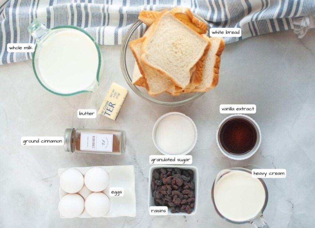 Ingredients for bread pudding arranged on a counter: whole milk, white bread, vanilla extract, granulated sugar, heavy cream, raisins, eggs, ground cinnamon, and butter.