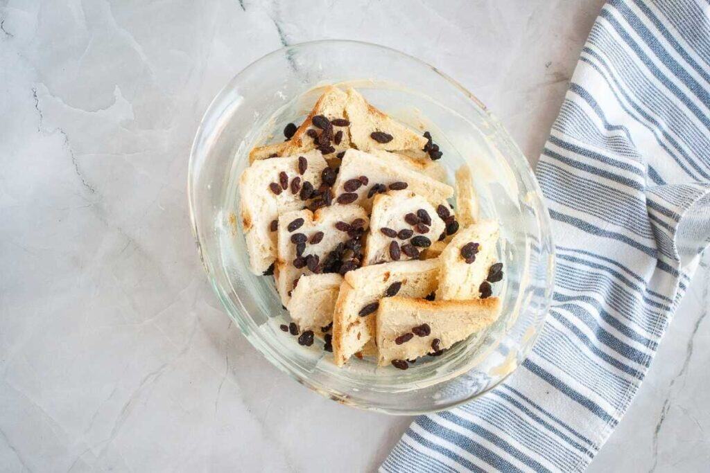 A glass bowl containing torn pieces of white bread and raisins sits on a marble surface next to a blue and white striped cloth.