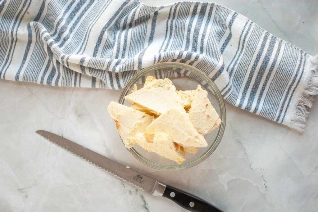 A glass bowl with sliced bread pieces sits on a marble surface next to a serrated knife and a folded blue-and-white striped cloth.