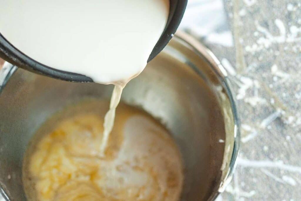 Milk is being poured from a black pot into a metal bowl containing a yellow mixture, with some flour scattered on the side.