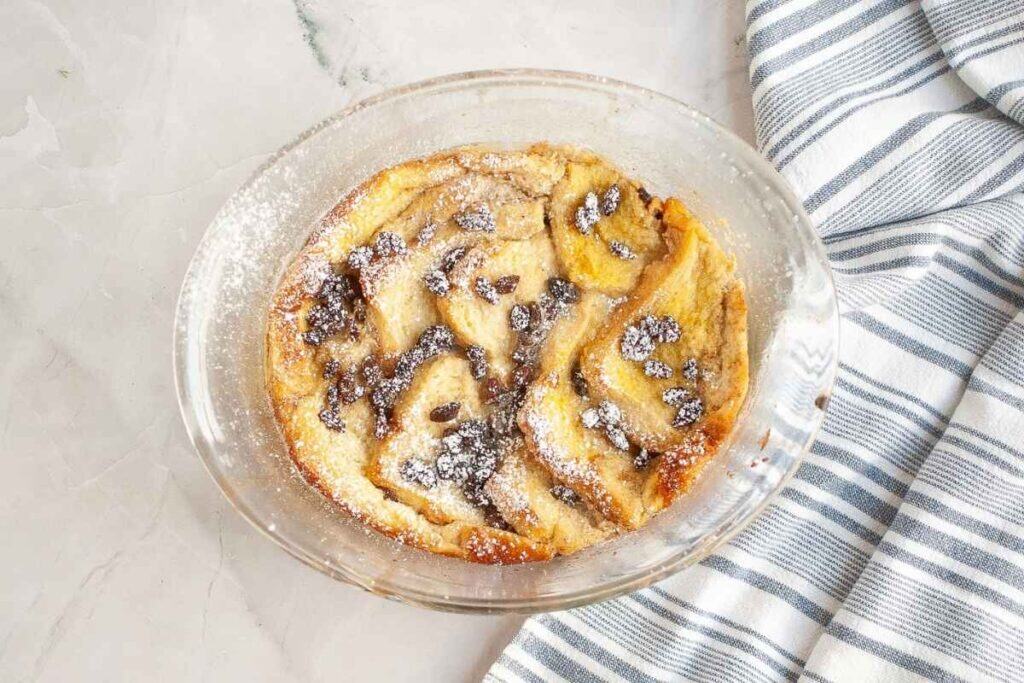 A round glass dish filled with baked bread pudding topped with powdered sugar sits on a marble surface next to a folded striped kitchen towel.