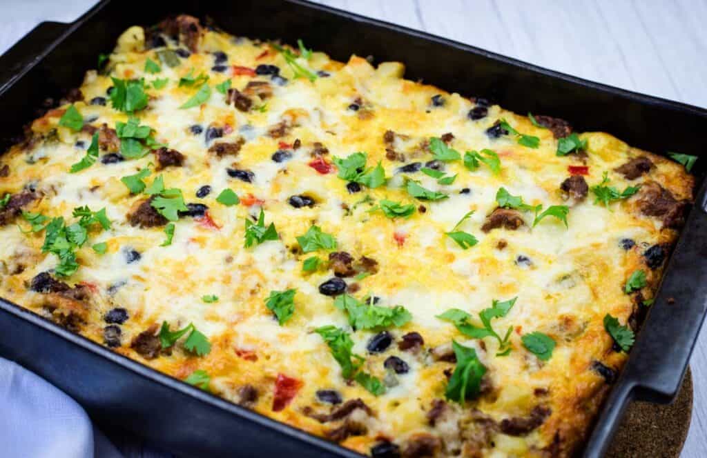 A baked casserole in a black rectangular dish, topped with melted cheese, black beans, ground meat, chopped red peppers, and fresh cilantro.