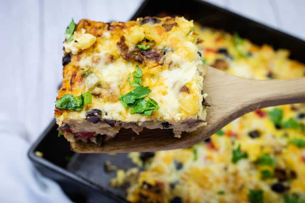 A wooden spatula lifts a square piece of baked casserole with cheese, vegetables, and herbs, over a baking dish.