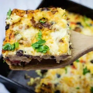 A wooden spatula lifts a square piece of baked casserole with cheese, vegetables, and herbs, over a baking dish.