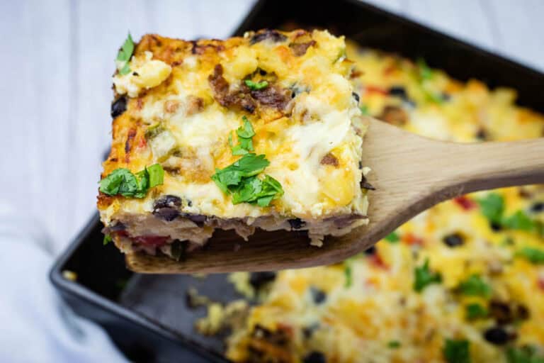A wooden spatula lifts a square piece of baked casserole with cheese, vegetables, and herbs, over a baking dish.