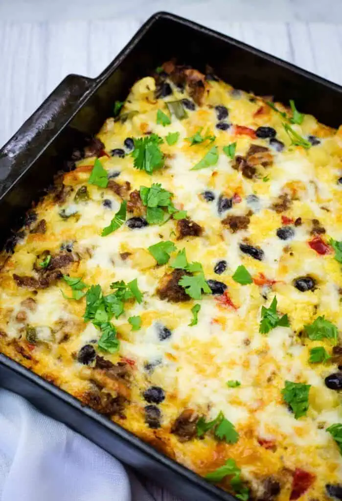 A baked breakfast casserole in a square pan, topped with melted cheese, ground meat, black beans, chopped vegetables, and fresh cilantro.