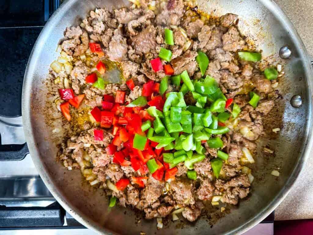 Ground beef cooking in a skillet with chopped green and red bell peppers and onions on a stovetop.