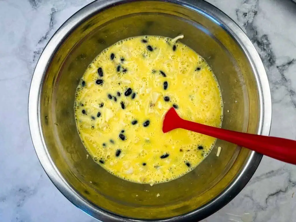 A metal bowl filled with a mixture of beaten eggs, black beans, and shredded cheese, with a red spatula resting in the bowl on a marble surface.