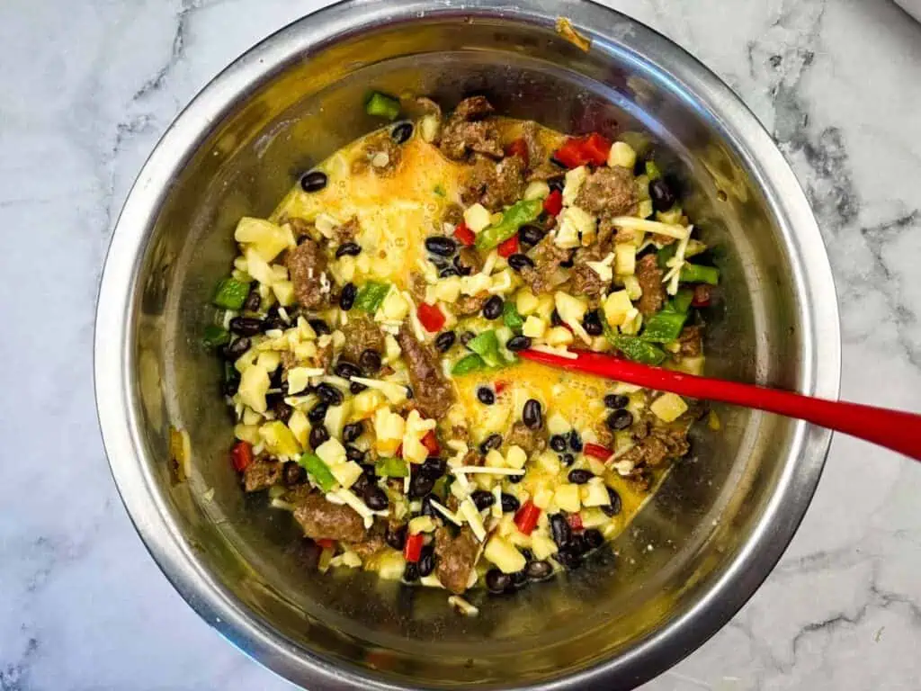 A stainless steel bowl containing a mixture of chopped vegetables, black beans, cheese, and pieces of meat with a red spoon on a marble surface.