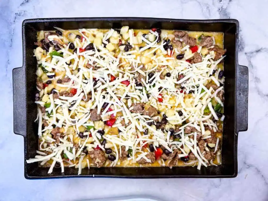 Rectangular baking dish filled with a casserole mixture of shredded cheese, beans, vegetables, and pieces of meat, on a marble countertop.