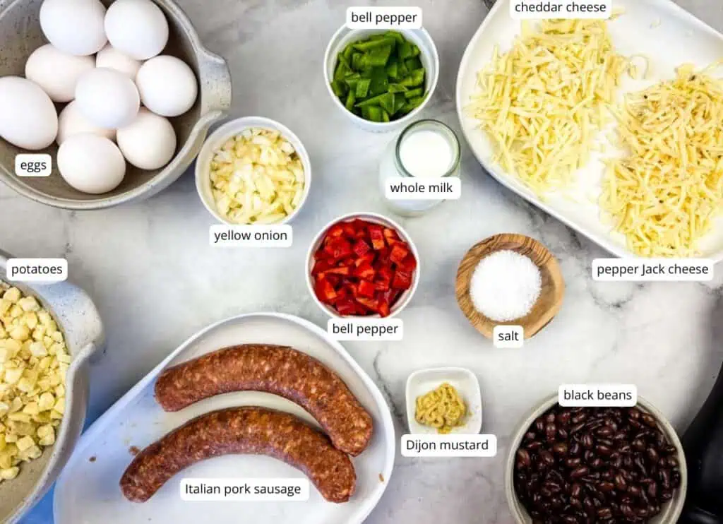 Ingredients for a breakfast casserole are arranged on a countertop, including eggs, potatoes, sausage, cheese, peppers, onion, black beans, milk, salt, and Dijon mustard.