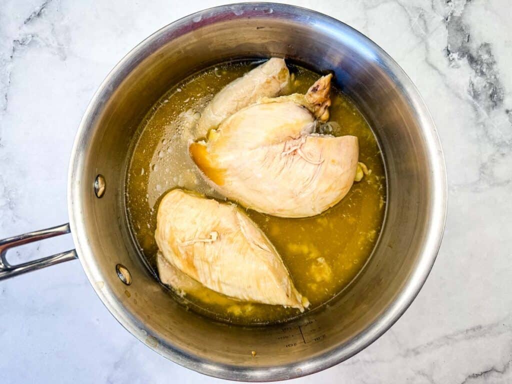 Cooked chicken breasts in broth inside a stainless steel pot, placed on a white marble surface.