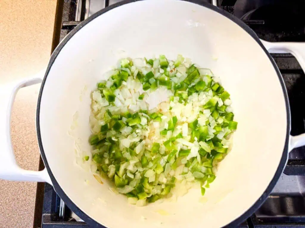 Chopped onions and green bell peppers sautéing in a white pot on a stovetop.