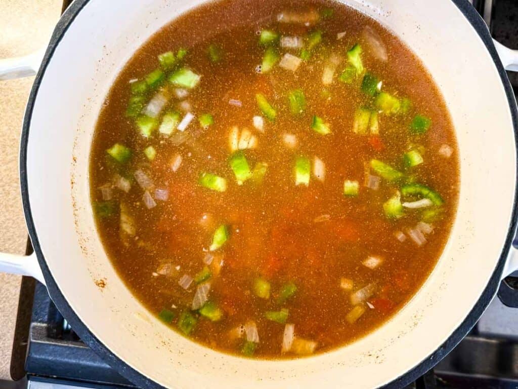 A white pot on a stove contains a simmering broth with chopped green bell peppers and onions.