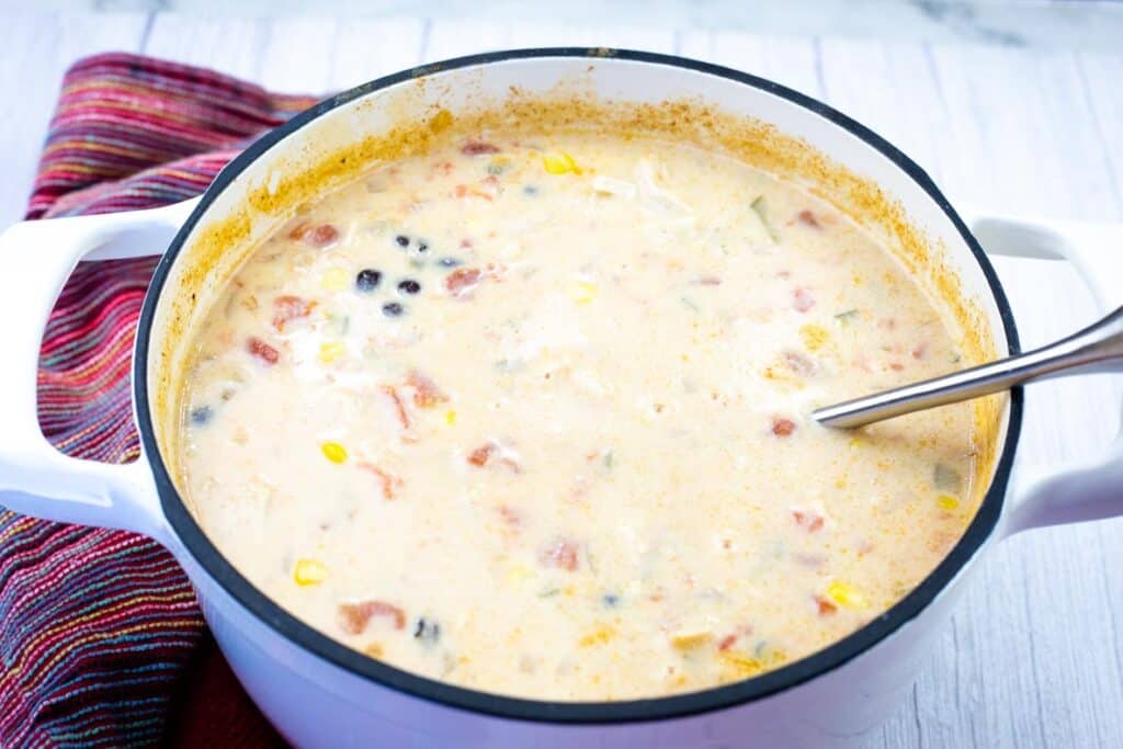 A white pot filled with creamy soup containing black beans, corn, tomatoes, and other ingredients, with a metal ladle inside.