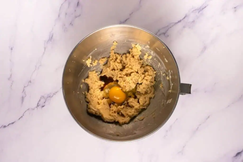 A metal mixing bowl containing partially mixed cookie dough, a raw egg, and some vanilla extract on a marble surface.