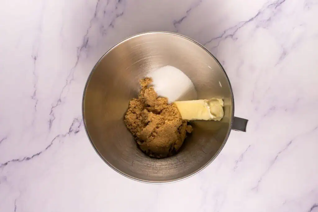 A metal mixing bowl contains brown sugar, white sugar, and a stick of butter on a marble countertop.