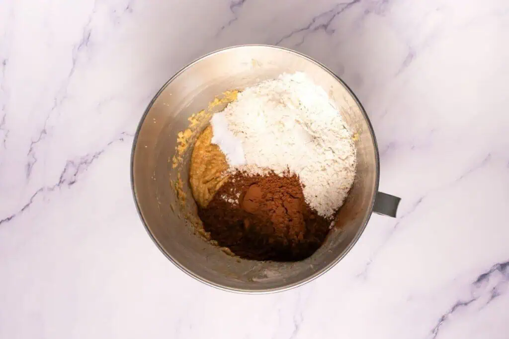 A mixing bowl containing flour, cocoa powder, and a wet batter mixture, viewed from above on a white marble surface.