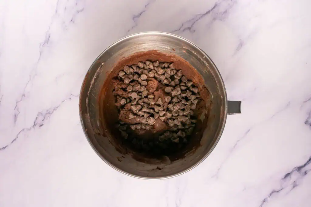 A metal mixing bowl containing chocolate batter with chocolate chips on top, placed on a white marble surface.