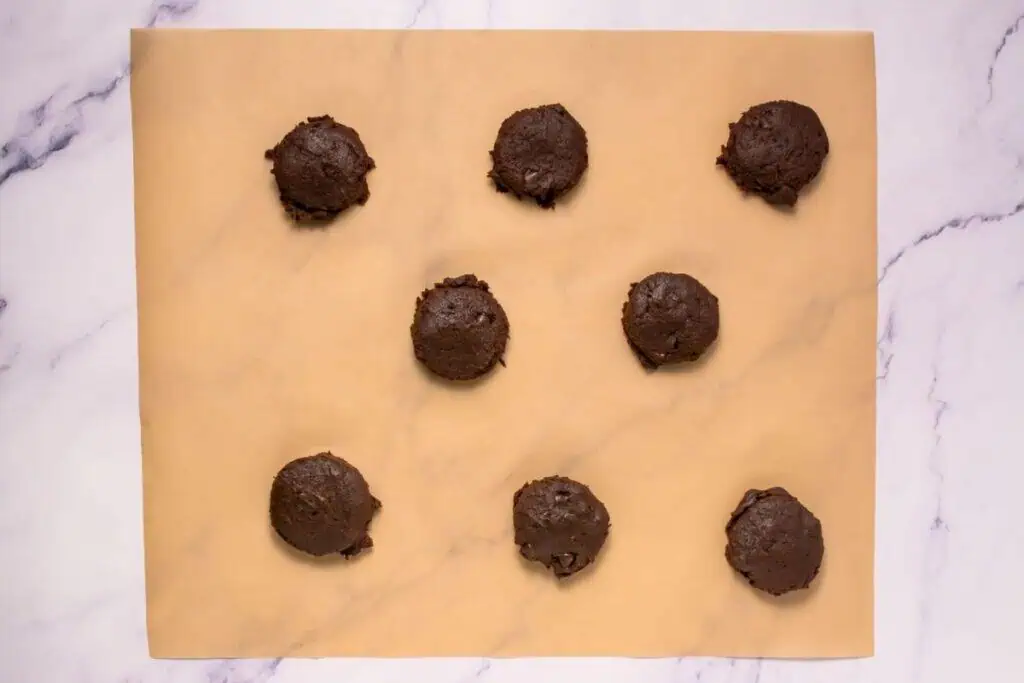 Eight scoops of chocolate cookie dough arranged in rows on a sheet of parchment paper on a marble surface.
