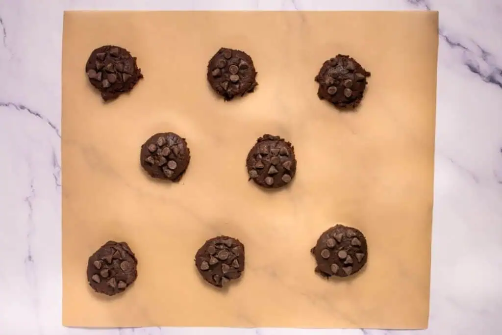 Nine unbaked chocolate chip cookie dough balls spaced evenly on a parchment-lined baking sheet, viewed from above.