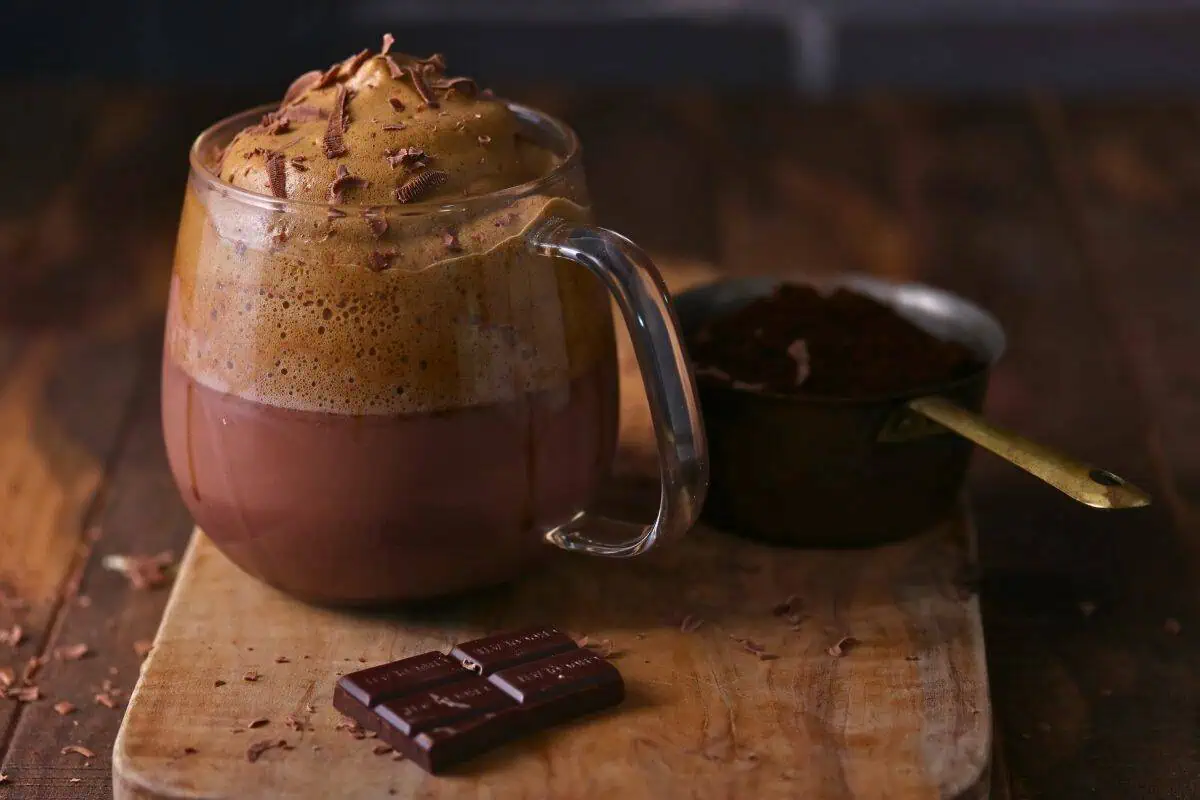 A glass mug of Dalgona Mocha Latte sits on a wooden board next to a brass cup of cocoa powder and pieces of dark chocolate.