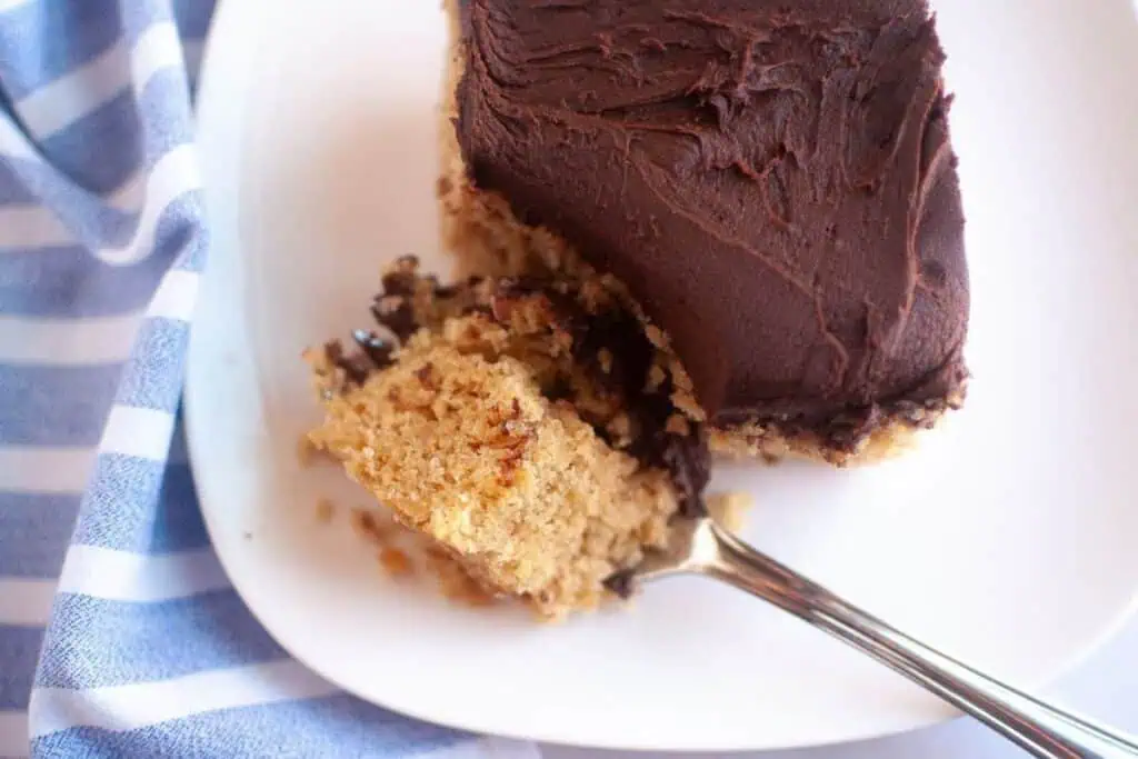 A square piece of cake with chocolate frosting on a white plate, with a fork holding a bite-sized piece.