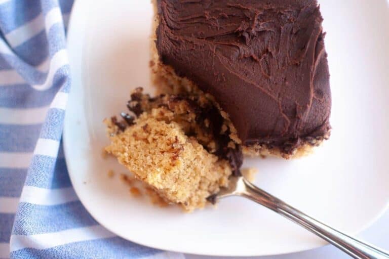 A square piece of cake with chocolate frosting on a white plate, with a fork holding a bite-sized piece.