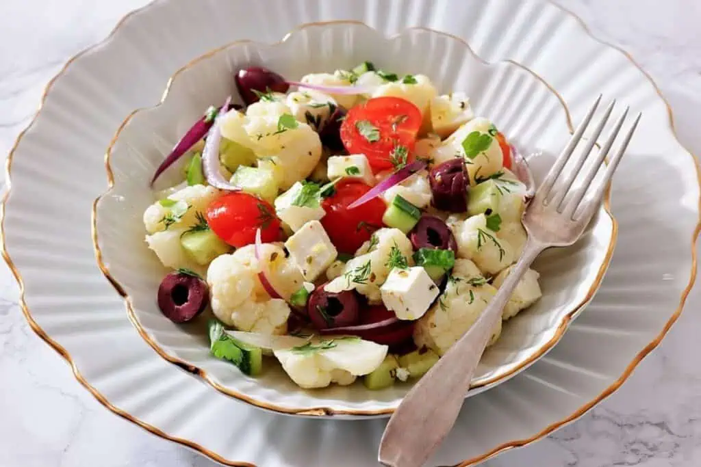 A plate of cauliflower salad with cherry tomatoes, cucumber, red onion, feta cheese, black olives, and herbs, served with a fork.