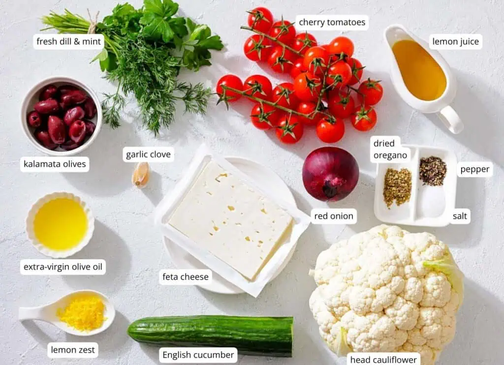 Assorted ingredients for a Mediterranean salad, including cauliflower, cucumber, feta cheese, kalamata olives, herbs, cherry tomatoes, red onion, and seasonings, displayed on a white surface.